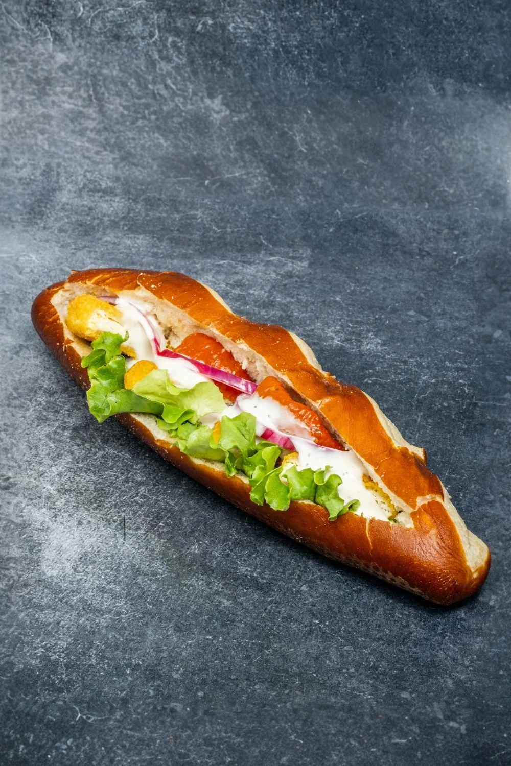 Sandwich au pain bretzel garni de chèvre chaud sur un fond gris. Sandwich réalisé par la boulangerie Maison Millet.